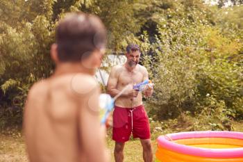 Father And Son Wearing Swimming Costumes Having Water Fight With Water Pistols In Summer Garden