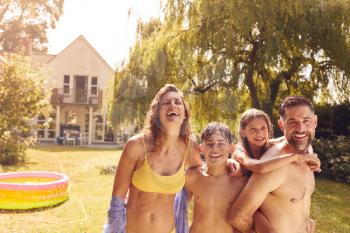 Portrait Of Family Wearing Swimming Costumes Having Fun Playing In Water From Garden Sprinkler