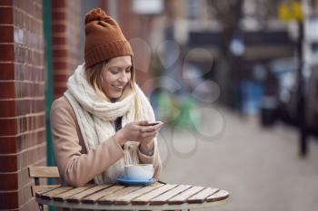 Female Customer Sending Text Message Sitting Outside Coffee Shop On Busy City High Street In Fall
