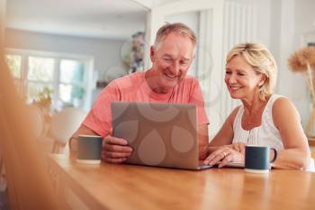 Retired Couple At Home In Kitchen Using Laptop To Shop Online Or Make Video Call