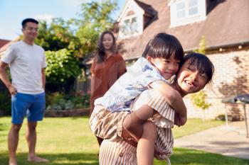 Asian Family Having Fun In Summer Garden At Home With Children Giving Piggyback Rides To Each Other