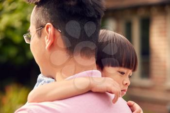 Loving Asian Father Cuddling Son In Garden As Boy Looks Over His Shoulder