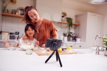 Asian Mother And Daughter Baking Cupcakes In Kitchen At Home Whilst On Vlogging On Mobile Phone