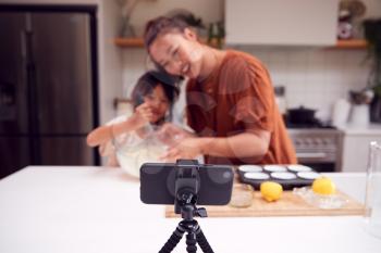 Asian Mother And Daughter Baking Cupcakes In Kitchen At Home Whilst On Vlogging On Mobile Phone