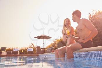 Loving Couple On Summer Vacation Sitting On Edge Of Swimming Pool With Cold Drinks