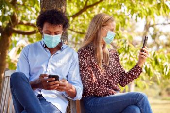 Couple Wearing Masks Meeting In Outdoor Park During Health Pandemic Looking At Mobile Phones