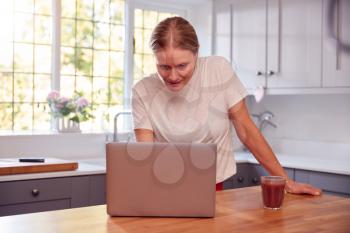 Mature Woman Wearing Fitness Clothing At Home In Kitchen Logging Activity On Laptop