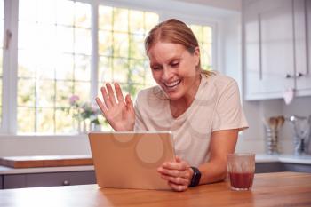 Mature Woman At Home In Kitchen Waving As She Makes Video Call On Digital Tablet