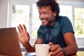 Mature Man At Home In Kitchen Waving As He Makes Video Call On Laptop Computer