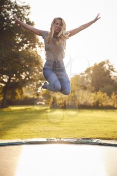 Portrait Of Woman In Mid Air Jumping On Trampoline Against Flaring Sun In Garden