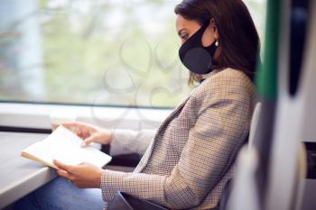 Businesswoman On Train Relaxing And Reading Book Wearing PPE Face Mask During Health Pandemic