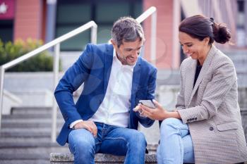 Businessman And Businesswoman Sitting By Steps Having Meeting Outdoors Looking At Phone