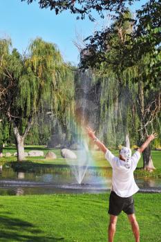 The tourist in a white easy shirt admires beauty of a landscape. Decorative fountain and shining rainbow on the golf course