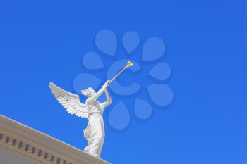 A white statue of a winged troubadour against the blue sky. The statue is installed on the roof eaves