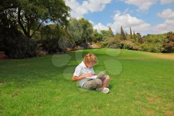 A beautiful blond boy plays on the laptop on a lawn in the city park