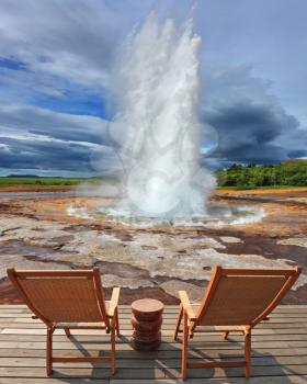  Geyser Strokkur in Iceland. Pillar of hot water and steam from forcing its way out of the ground. Two lounge chairs and  small table on  wooden platform for easy observation
