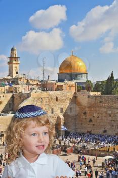 The Jewish holiday of Sukkot. Cute little boy with long blond curls and blue eyes in knitted skullcap. He stands at the main Jewish shrine - Western Wall of Temple