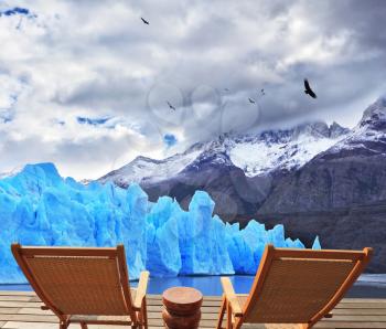 Two folding wooden chairs and a small bedside table on the boardwalk. A comfortable place to enjoy the beauty of the enormous Perito Moreno glacier in Argentina.