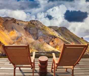 Two comfortable wooden chairs and small table stand for tired tourists.  The rhyolite mountains in volcanic summer tundra. Travel to Iceland in July