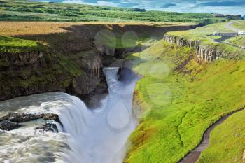 Grandiose Gullfoss in Iceland. In July raging water is shined with the bright morning sun. River banks grew with a green northern moss