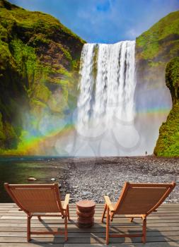 Magnificent powerful jet Skógar River forms a large rainbow. The famous waterfall Skógafoss, Iceland. On stony ground in front of the waterfall are two wooden deck chairs
