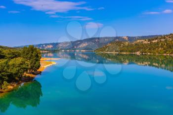 Smooth emerald green water of Lake Sainte-Croix-du-Verdon reflects the sky and wooded shore. Mountain canyon Verdon in the French Alps