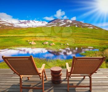 At the lake on wooden platform there are two deckchairs. In the smooth water of lake reflects cold cloudy sky