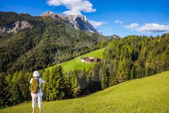  Forested mountains surrounded by green Alpine meadows. Active elderly woman-tourist with backpack photographs at Dolomites, Northern Italy. The concept of an active and eco-tourism
