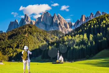  Dolomites, Tirol. Active woman-tourist with backpack photographs the church of Santa Maddalena. Forested mountains surrounded by green Alpine meadows. The concept of ecological and active tourism