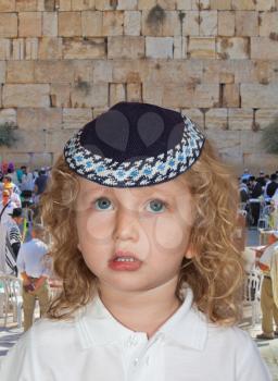  The Jewish holiday of Sukkot. Cute little boy with long blond curls and blue eyes in knitted skullcap. He stands at the main Jewish shrine - Western Wall of Temple
