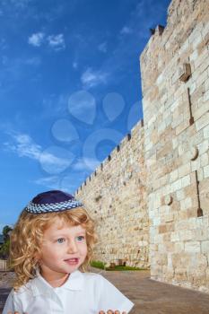 The Jewish holiday of Sukkot. Cute little boy with long blond curls and blue eyes in a knitted skullcap. He stands at the fortress walls of the Old City of Jerusalem