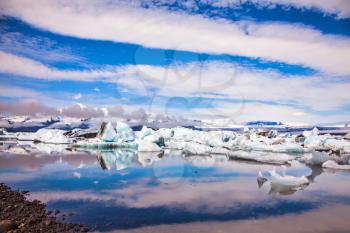 The procession of luminous icebergs on Jokulsarlon, Iceland. The concept of extreme northern tourism. Striped clouds beautifully reflected in the smooth water of lagoon