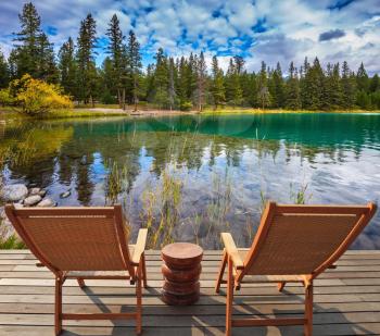 Pretty round lake in the coniferous forest. Two comfortable lounge chairs on wooden pedestal on the lake. Canadian Rocky Mountains, Jasper National Park