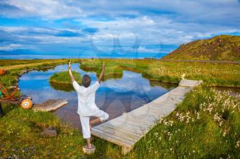Woman in white performs yoga. Iceland. Small pond with thermal water. In the smooth surface of the water reflects the sky and clouds