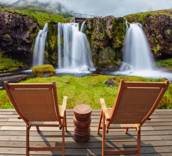 Deck chairs on wooden platform waiting for tourists.  Threaded full-flowing waterfall Kirkyufell Foss on the grassy mountains