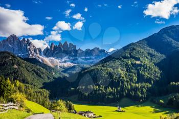 Picturesque little church of Santa Maddalena in valley Val di Funes. Rocky peaks and forested mountains surrounded by green Alpine meadows. Sunny day in Dolomites, Tirol