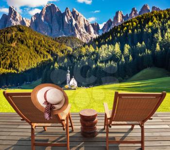 Two deck chairs on a wooden floor installed to relax and review the Alpsg and church of Santa Magdalena. The concept of eco-tourism in Alpine meadows