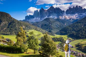 Woman-tourist with backpack photographs at Dolomites, Northern Italy. Forested mountains surrounded by green Alpine meadows and farm. The concept of active and eco-tourism