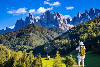 Active elderly woman-tourist with backpack photographs the church of Santa Maddalena in Dolomites. Forested mountains surrounded by green Alpine meadows. The concept of ecological and active tourism