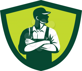 Illustration of an organic farmer wearing hat and overalls arms folded looking to the side viewed from front set inside shield crest on isolated background done in retro style. 