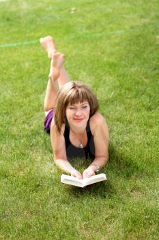 beautiful young female student reading a book outdoors laying on grass