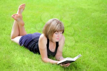 Beautiful young smiling woman laying on grass reading book, against green of summer park.