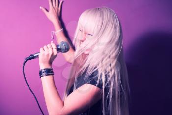 Blonde woman in karaoke sing with microphone on pink backgound. She is in motion and her lond hair shaking around her head.