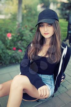 beautiful teenage lady with long legs in jeans short sits on skateboard in park setting. Her long silky hair on shoulders, she is looking at camera