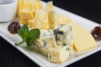 Cheese plate on a dark background table. Many kinds of cheese with sauce and greens on a white plate, closeup shot