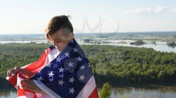 Blonde boy waving national USA flag outdoors over blue sky at the river bank. Beauty summer sunny day. American flag, patriotism, independence day 4th july concept