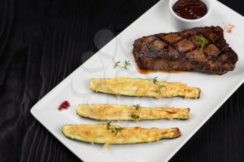 Grilled black angus strip loin steak New York with zucchini on white plate on black wooden background with