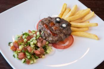 Funny meat cutlet face with french fries as hair and cutted vegetables for children menu
