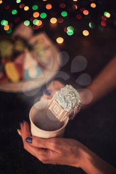Female hands with cup of tea and christmas cookies house on dark color bokeh lights background