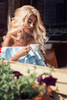 Beauty sensual woman in elegant blue dress in cafe. Young woman sitting in cafe with cup of coffee in summer day.